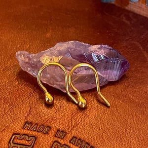 May include: Two gold-colored earring hooks with a small ball at the end of each hook. The hooks are resting on a purple crystal.