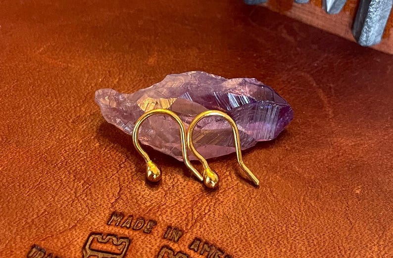 May include: Two gold-colored earring hooks with a small ball at the end of each hook. The hooks are resting on a purple crystal.