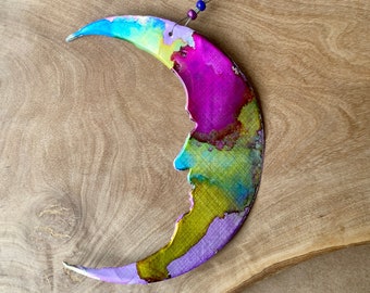 May include: A crescent moon shaped ornament with a colorful abstract design in shades of pink, purple, blue, green, and yellow. The ornament is made of metal and has a wire hanger.