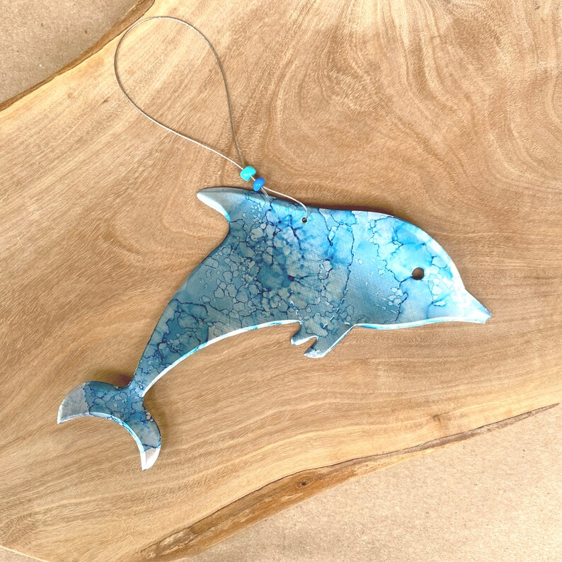 May include: A blue and silver dolphin ornament with a marbled effect. The dolphin is hanging from a thin silver string with blue beads.