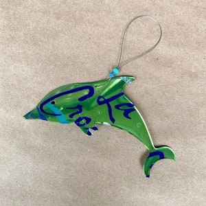 May include: A green, blue, and white dolphin ornament made from a recycled aluminum can. The dolphin is hanging from a silver wire with a blue bead. The can has the text "La Croix" printed on it.