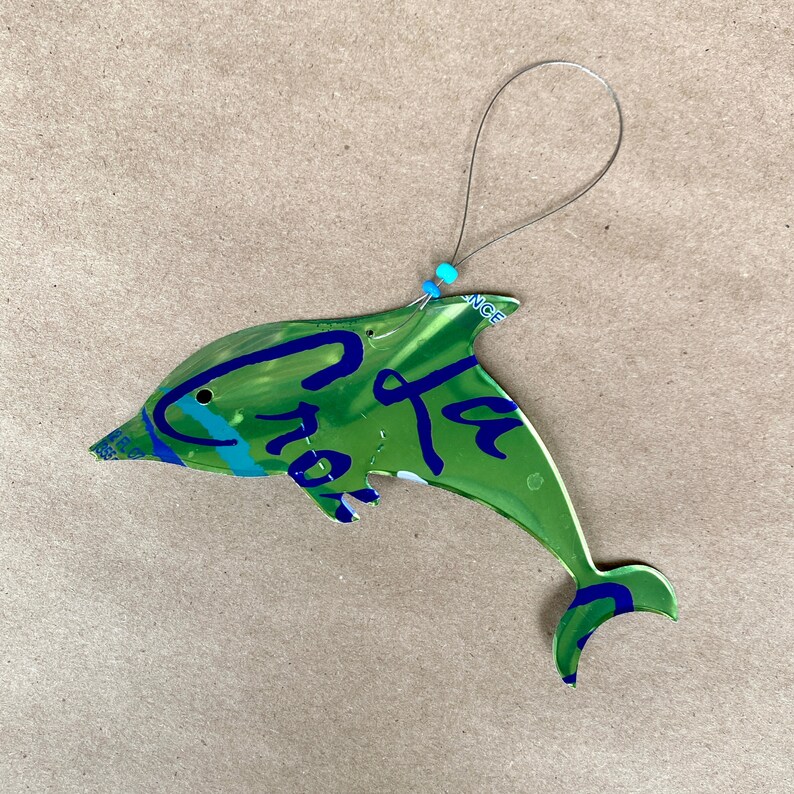 May include: A green, blue, and white dolphin ornament made from a recycled aluminum can. The dolphin is hanging from a silver wire with a blue bead. The can has the text "La Croix" printed on it.