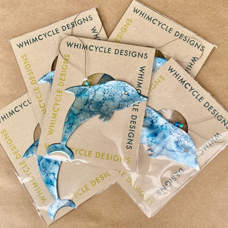 May include: Five blue and white dolphin ornaments with a marbled effect, packaged on brown cards with the text "Whimsycycle Designs" printed on them.