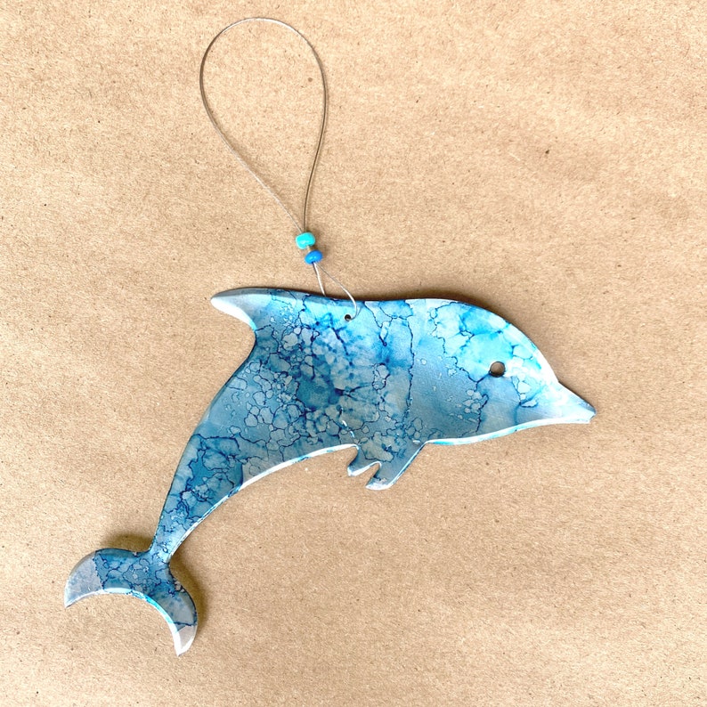 May include: A blue and white dolphin ornament with a marbled finish. The dolphin is suspended from a silver string with two blue beads.