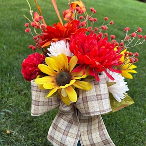 May include: A colourful floral display with red, yellow, and white blooms, complemented by orange and red berries. A plaid ribbon bow provides a rustic feel. The bouquet is set against a backdrop of green grass.