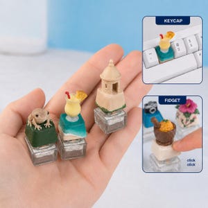 May include: Three miniature keycaps featuring a small frog on a green base, a tropical drink on a beach-themed base, and a tan stone tower. Additional images show a keycap on a keyboard and a fidget toy with a mortar and pestle design.