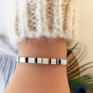 Peut inclure: Un bracelet en perles blanches, noires et dorées avec un fermoir doré. Le bracelet est porté au poignet.