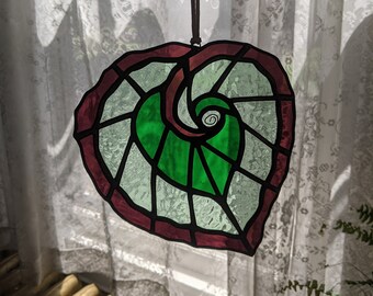 Puede incluir: Una ventana de vitral colgada con un diseño de hoja roja y verde. La hoja tiene un patrón en espiral en el centro.