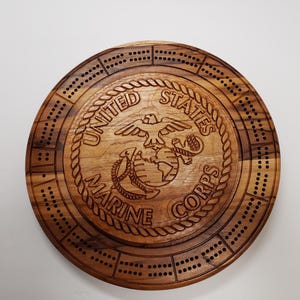 May include: A wooden cribbage board with a United States Marine Corps logo in the center. The board is circular and has a dark brown wood grain.