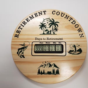 May include: A wooden circular countdown clock with a digital display that reads "999 07 07 03". The clock is decorated with images of palm trees, a fishing scene, a golfer, and mountains. The text "Retirement Countdown" is printed around the top of the clock. The text "Days to Retirement" is printed below the digital display.