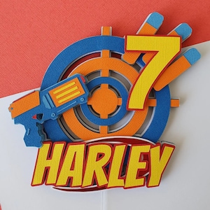 May include: A layered paper cake topper with a blue and orange toy gun, a target, and the number 7 in yellow. The name "HARLEY" is displayed in yellow with a red outline. The background is red and white.
