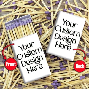 May include: Two white matchboxes with the text "Your Custom Design Here" in black. One box is open, revealing purple-tipped matches. The boxes are surrounded by scattered wooden matches with purple tips.