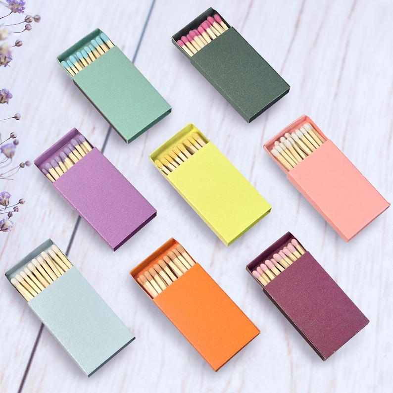 May include: Eight matchboxes with different colored covers. The matchboxes are arranged in a grid pattern on a white wooden surface. The matchboxes are all the same size and shape. The matchboxes are filled with matches. The colors of the matchboxes are green, purple, yellow, orange, pink, red, blue, and brown.