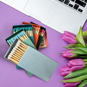 May include: A set of six matchboxes with different colored covers, including teal, green, orange, and black. The matchboxes are arranged in a fan shape on a purple surface. A bouquet of pink tulips is in the background.