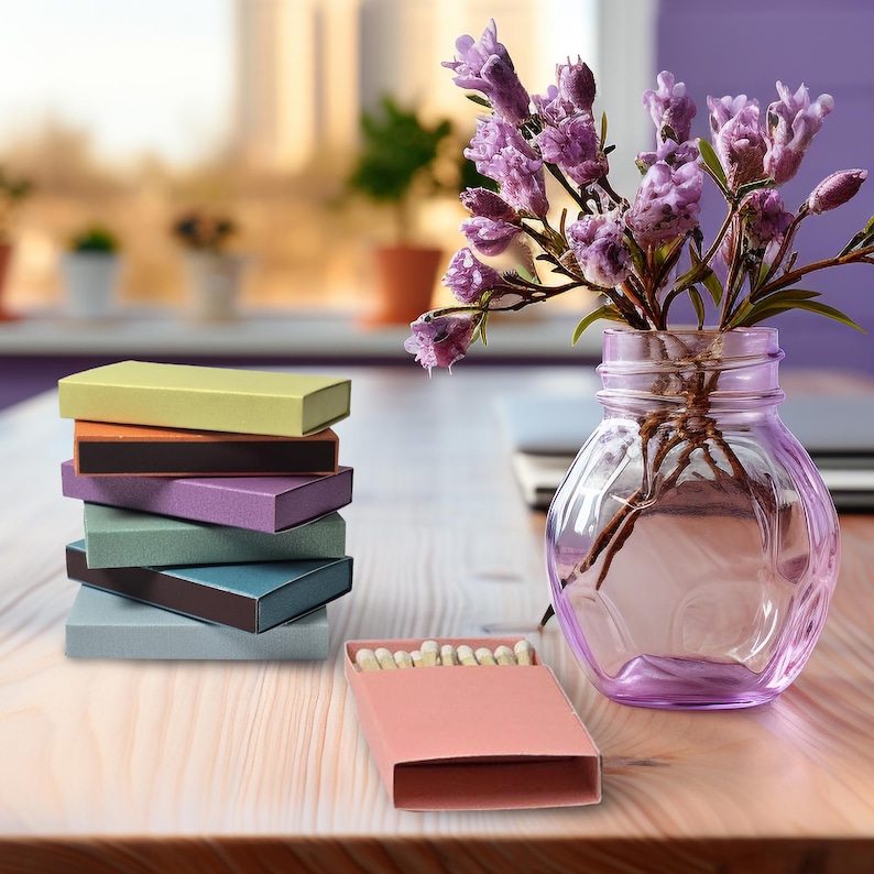May include: A stack of six colorful matchboxes with a pink matchbox in front. The matchbox is open and shows a row of matches. A vase with purple flowers sits on a wooden table.