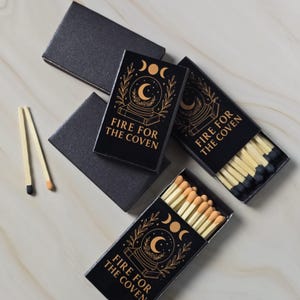 May include: Three black matchboxes with gold foil designs featuring a crystal ball, moon phases, and the text FIRE FOR THE COVEN. Two matchboxes are open, revealing wooden matches with black and tan tips. Two loose matches lie nearby.