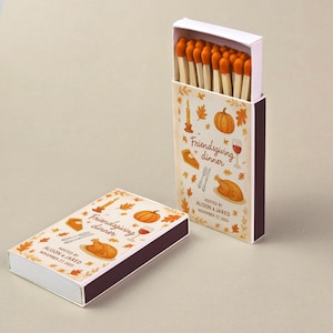 May include: Two matchboxes, one open, featuring a Thanksgiving theme. The boxes are white with a brown border and are decorated with illustrations of a pumpkin, turkey, and wine glass. Text on the box reads "Friendsgiving dinner" and "Hosted by Alison & Jared November 27, 2025."