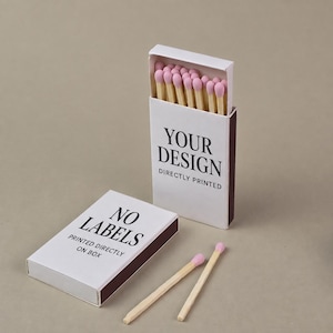 May include: A white matchbox with light pink tipped matches inside, featuring the black text YOUR DESIGN DIRECTLY PRINTED. A second closed white matchbox lies flat with the black text NO LABELS PRINTED DIRECTLY ON BOX, alongside two loose matches.