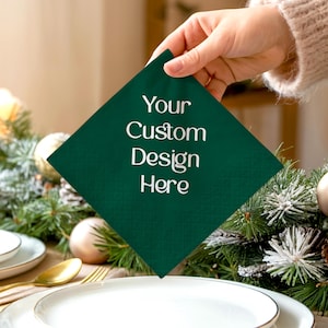 May include: A dark green square napkin with the text "Your Custom Design Here" in white. The napkin is held above a table setting with white plates, gold silverware, and holiday greenery.