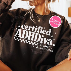 May include: A dark brown sweatshirt with the text "certified ADHDiva. dramatic & distracted" in white. A pink circle with the text "6 Colors Included" is on the right side of the sweatshirt.