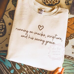May include: Cream-colored sweatshirt with the text "running on snacks, scripture, and his saving grace" in a handwritten font, with a heart symbol. The sweatshirt has a crew neck and long sleeves. The design includes the text "Ava Rose DIGITAL DESIGN".