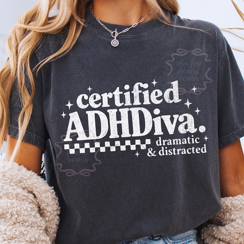 Pode incluir: T-shirt cinzento escuro com o texto branco "certified ADHDiva. dramatic & distracted". A camisola tem um aspeto envelhecido e um padr&atilde;o xadrez por baixo do texto.