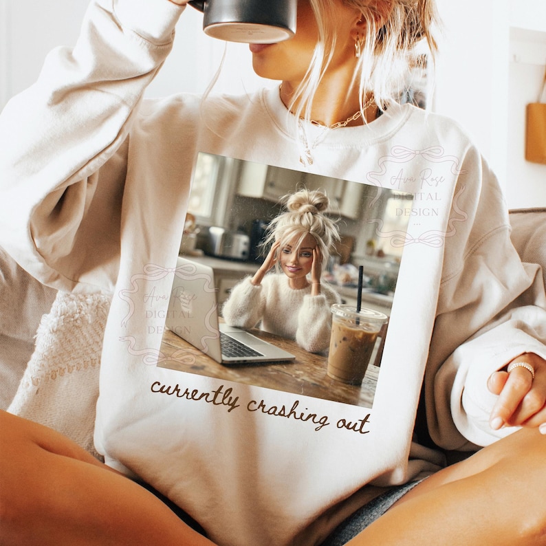 Peut inclure: Un sweat-shirt beige avec l'inscription "currently crashing out". Le sweat-shirt pr&eacute;sente un graphisme d'une poup&eacute;e assise devant un ordinateur portable avec un caf&eacute;. La poup&eacute;e porte un pull blanc.
