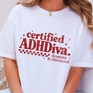 Pode incluir: Camiseta branca com o texto vermelho "certified ADHDiva. dramatic & distracted". O design inclui estrelas e um padr&atilde;o xadrez. Um colar de ouro com um pingente de cora&ccedil;&atilde;o &eacute; vis&iacute;vel.