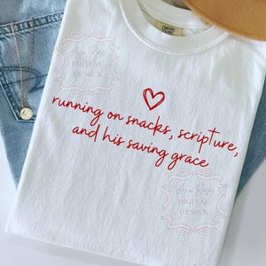 May include: White t-shirt with red text that reads "running on snacks, scripture, and his saving grace" with a red heart. The shirt is next to a pair of blue jeans and a gold necklace.