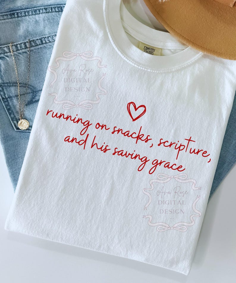 May include: White t-shirt with red text that reads "running on snacks, scripture, and his saving grace" with a red heart. The shirt is next to a pair of blue jeans and a gold necklace.