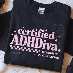 Pode incluir: Camisola preta com o texto "certified ADHDiva. dramatic & distracted" em rosa. A camisola tem gola redonda e mangas compridas. A marca "Gildan" &eacute; vis&iacute;vel na etiqueta.