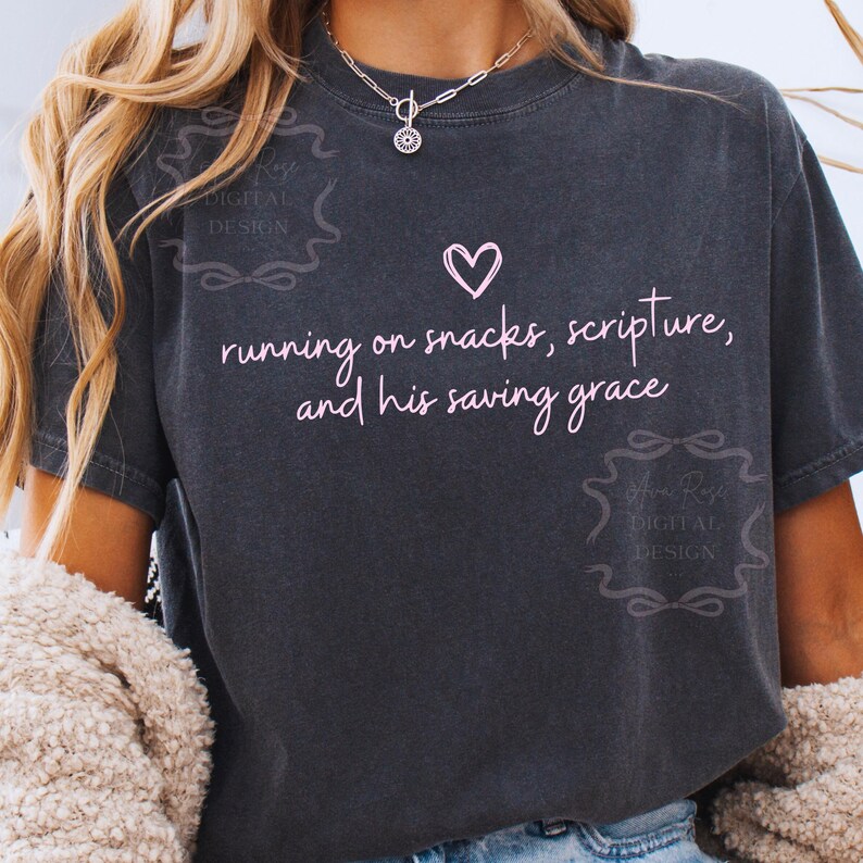 May include: Dark gray t-shirt with the text "running on snacks, scripture, and his saving grace" in pink script. A pink heart is above the text. The shirt is a casual style.