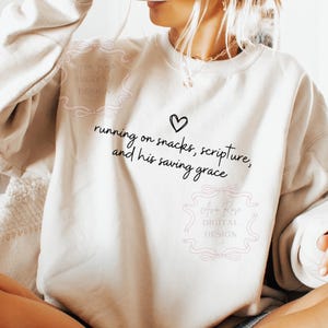 May include: Cream-colored sweatshirt with the text "running on snacks, scripture, and his saving grace" in black script, along with a heart symbol. The sweatshirt has a crew neck and long sleeves.