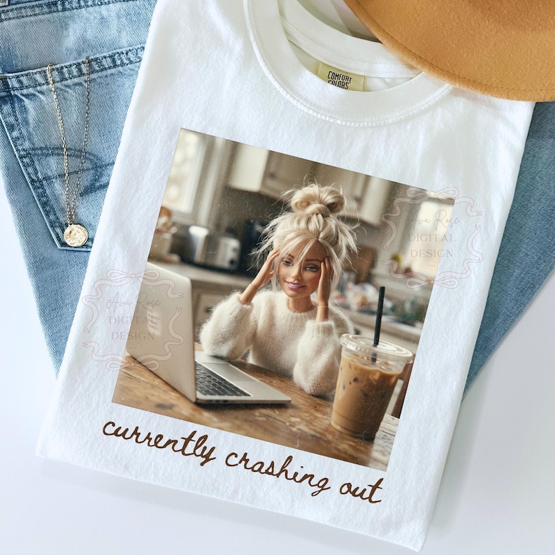 Peut inclure: T-shirt blanc avec un imprim&eacute; d'une poup&eacute;e utilisant un ordinateur portable, avec le texte marron "currently crashing out". La poup&eacute;e porte un pull blanc et a les cheveux blonds. Le t-shirt est sur un fond de jean bleu et d'un collier dor&eacute;.