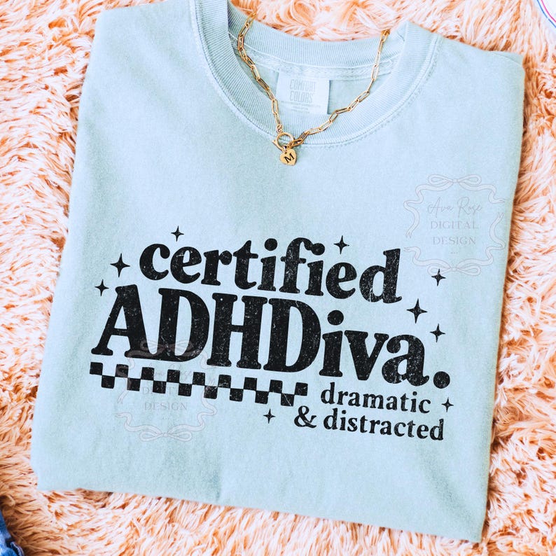 Pode incluir: Uma camisola azul clara com o texto "certified ADHDiva. dramatic & distracted" em preto. Um colar de corrente dourada com um pendente est&aacute; drapeado sobre a camisa. A camisa est&aacute; sobre uma superf&iacute;cie texturizada cor de p&ecirc;ssego.
