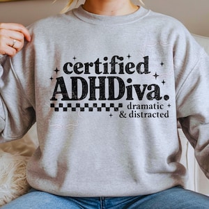 Pode incluir: Camisola cinza claro com a frase preta "certified ADHDiva. dramatic & distracted". O design inclui estrelas e um padr&atilde;o xadrez.