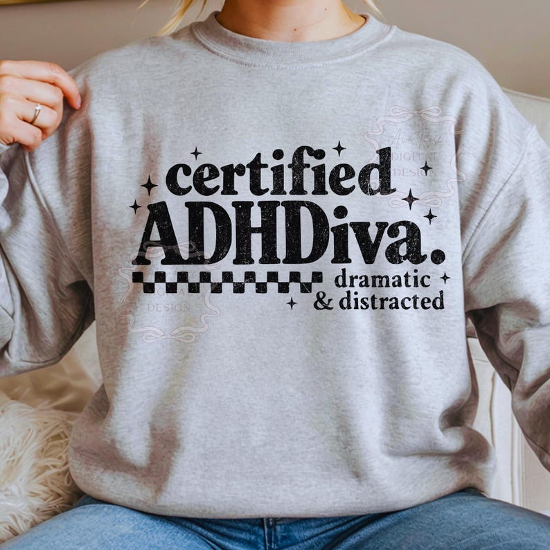 Pode incluir: Camisola cinza claro com a frase preta "certified ADHDiva. dramatic & distracted". O design inclui estrelas e um padr&atilde;o xadrez.