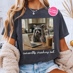 May include: Dark gray t-shirt featuring a raccoon with its hands on its head, looking stressed in front of a laptop and iced coffee. The text "currently crashing out" is printed below the image. A pink circle with "3 Colors Included" is in the upper right.
