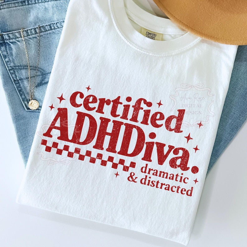 Pode incluir: Camiseta branca com o texto vermelho "certified ADHDiva. dramatic & distracted". A camiseta est&aacute; dobrada sobre jeans azuis, com um colar dourado e um chap&eacute;u marrom ao fundo.