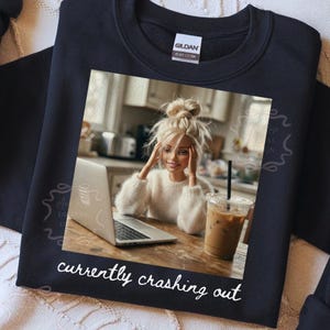Peut inclure: Sweat-shirt noir avec une image imprim&eacute;e d'une poup&eacute;e avec un ordinateur portable et un caf&eacute; glac&eacute;. L'image comprend le texte "currently crashing out" en &eacute;criture blanche. La poup&eacute;e porte un pull blanc.