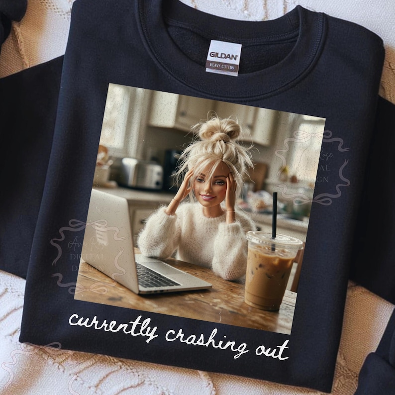 Peut inclure: Sweat-shirt noir avec une image imprim&eacute;e d'une poup&eacute;e avec un ordinateur portable et un caf&eacute; glac&eacute;. L'image comprend le texte "currently crashing out" en &eacute;criture blanche. La poup&eacute;e porte un pull blanc.