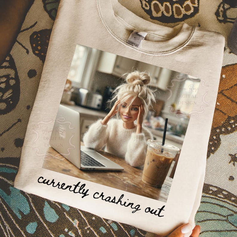 Peut inclure: Sweat-shirt cr&egrave;me avec une image imprim&eacute;e d'une poup&eacute;e assise &agrave; une table avec un ordinateur portable et un caf&eacute; glac&eacute;. L'image contient le texte "currently crashing out" en &eacute;criture manuscrite.