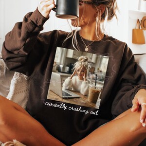 Peut inclure: Un sweat-shirt marron avec une image imprim&eacute;e d'une femme avec un ordinateur portable et un caf&eacute; glac&eacute;. Le texte "currently crashing out" est imprim&eacute; en dessous. La personne tient une tasse noire.