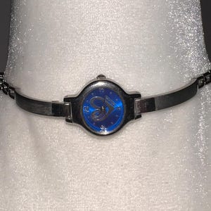 Puede incluir: Un reloj de pulsera plateado con una esfera azul y un diseño de corazón. El reloj tiene una pulsera plateada.