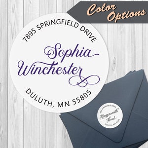 May include: A white round return address label with black text. The text reads "7895 Springfield Drive, Sophia Winchester, Duluth, MN 55805". The name "Sophia Winchester" is written in a cursive font.