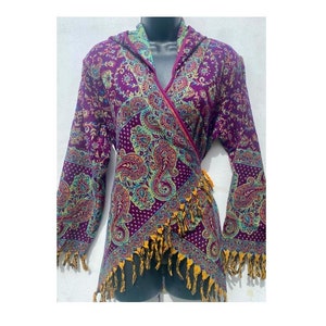 Puede incluir: Un top envolvente morado con un estampado de cachemira en verde azulado, amarillo y rosa. El envoltorio tiene mangas largas y un dobladillo con flecos. La prenda se exhibe en un maniquí.