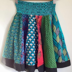 Op de afbeelding: Een kleurrijke rok met een turquoise elastische tailleband en panelen met verschillende patronen. De panelen tonen ontwerpen in turquoise, blauw, groen en rood, met een zwarte zoom. De rok hangt aan een houten kleerhanger.