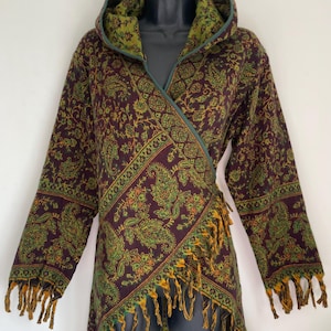 Könnte beinhalten: Ein Kapuzenwickel mit einem Paisley-Muster in Burgunder-, Grün- und Goldtönen. Der Wickel hat lange Ärmel und ist mit grünen Kanten und goldenen Fransen versehen. Der Wickel wird auf einer schwarzen Schaufensterpuppe präsentiert.