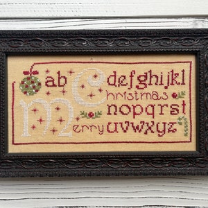 May include: A framed cross-stitch sampler with the alphabet and the word "christmas" in red embroidery thread. The sampler is on a white wood background.
