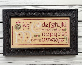 May include: A framed cross-stitch sampler with the alphabet and the word "christmas" in red embroidery thread. The sampler is on a white wood background.
